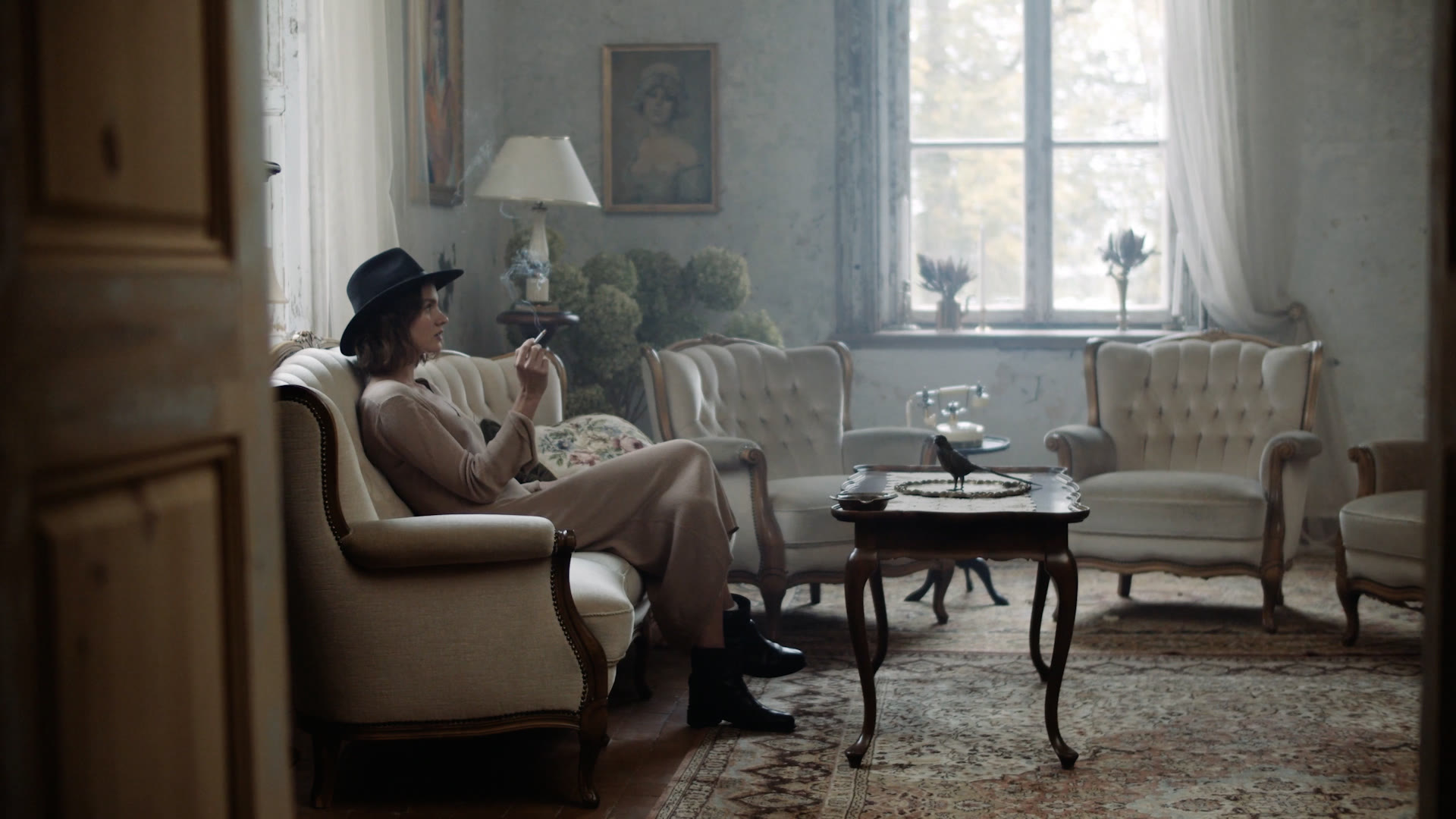 A woman in a hat smoking on a vintage sofa in a sunlit manor room, Playing with Ambiguity music video for Luisa Babarro
